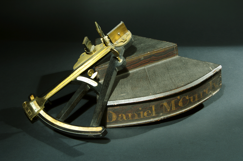 A historical navigational instrument for measuring angles up to 45 degrees. It has an arc that is one-eighth of a circle, hence the name “octant.” It has a wooden plinth with the name Daniel McCurdy written on it in gold. The instrument itself is made of a wooden a frame with brass fittings.