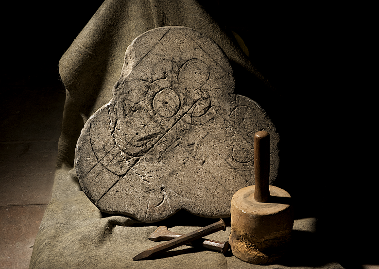 A posed photo of a mason's carving and tools. It looks almost like a still life. Asection of stone that looks like a trefoil of three leaf clover is carved with some rudimentary shapes, including circles and other trefoils. In front of it are some chisels and a masons hammer or mallet.