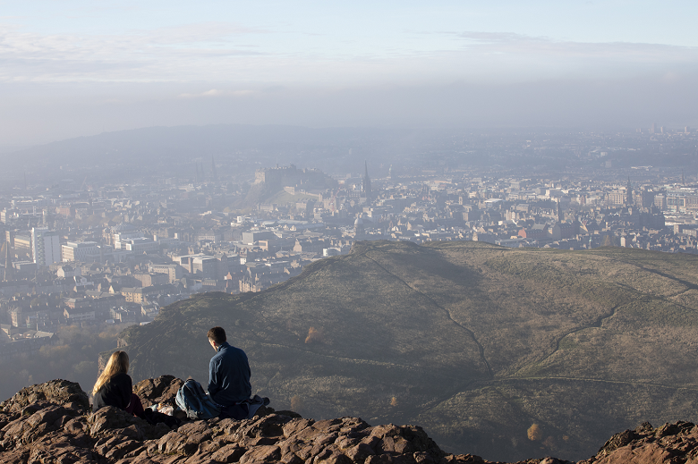 Two hikers sitting on a rocky ledge at the top of Arthur's Seat. They are looking down on the city of Edinburgh, with the castle in the centre of the view. 