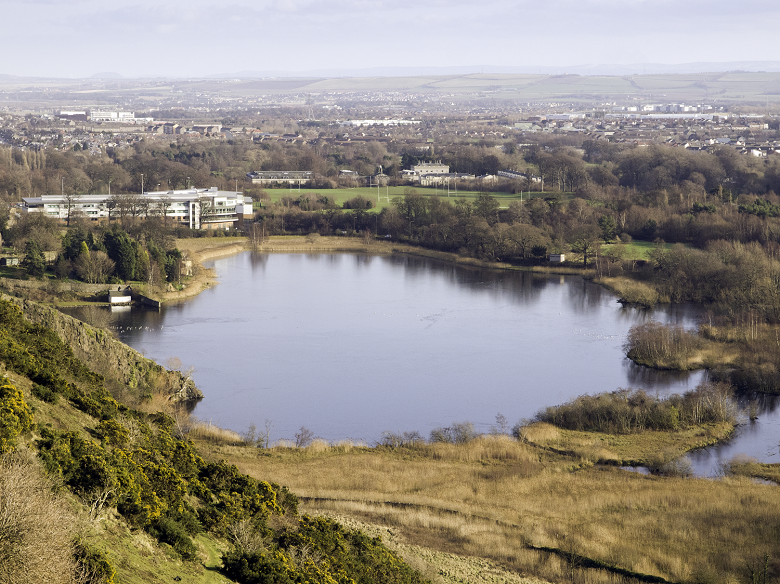 A view across a small loch towards suburban houses and a school building. 