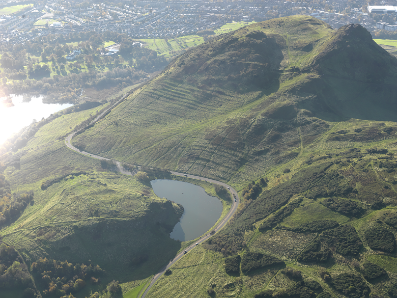 An aerial photo of a small loch surrounded by hills which have been shaped by Holyrood Park's archaeology. On the slopes there are lines and patterns caused by agricultural activity. 