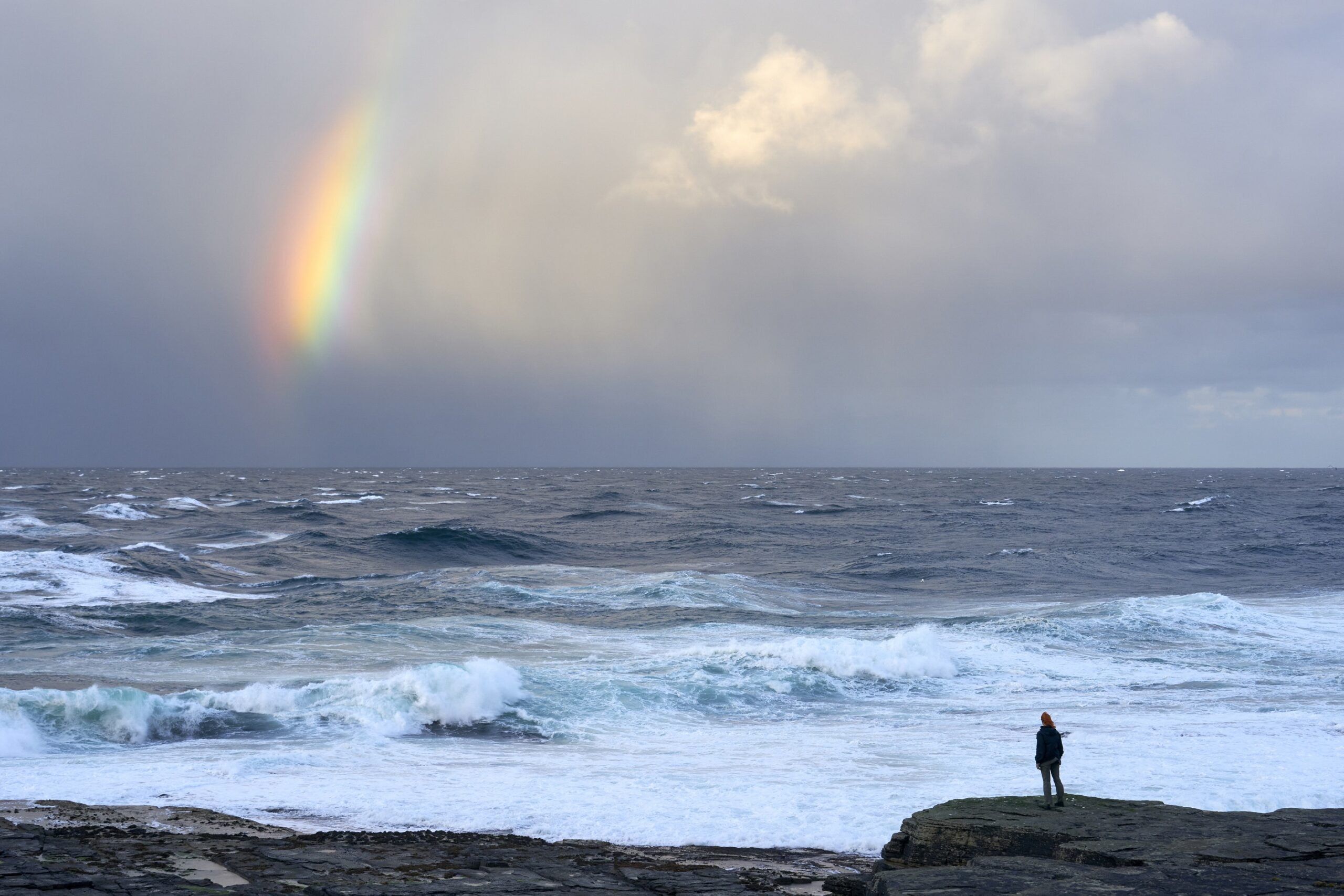 Film still from The Outrun showing Saoirse Ronan standing on the Orcadian shore overlooking the rough sea. There is a rainbow.