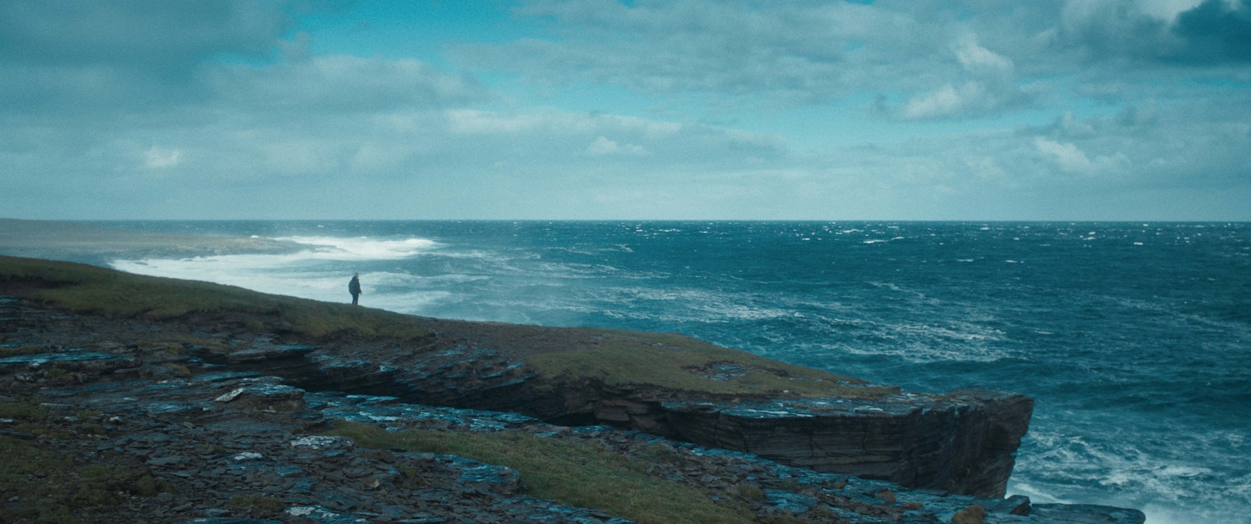 Film still from The Outrun showing Saoirse Ronan standing on the Orcadian shore overlooking on a dark and gloomy day.