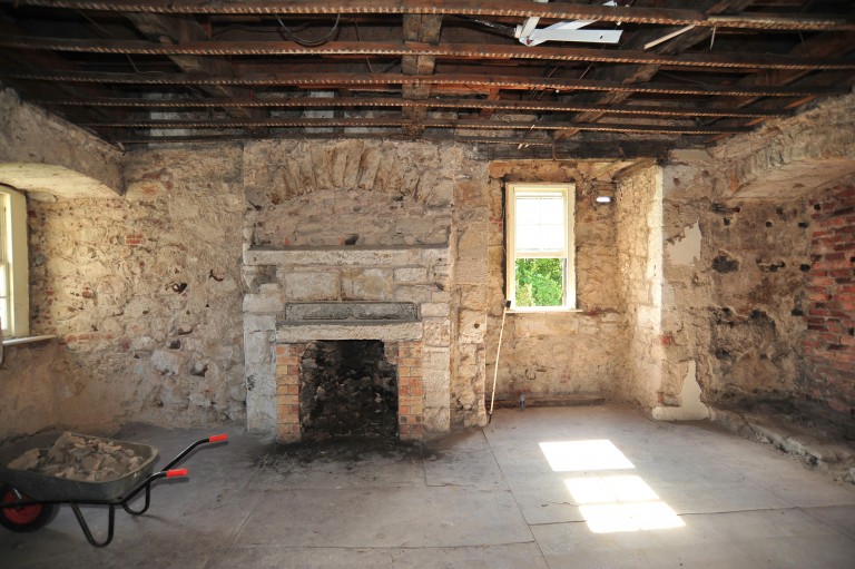 A crumbling stone fireplace next to a bright window. There is also a wheelbarrow filled with rubble.