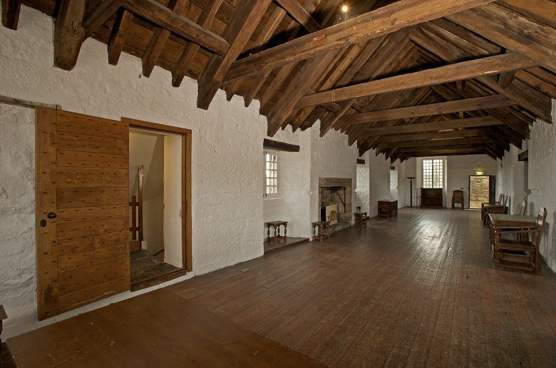 A large gallery inside Aberdour Castle. It has a wooden floor and a roof made from large wooden beams. Various pieces of historic furniture have been placed at intervals along the long, corridor-like room.