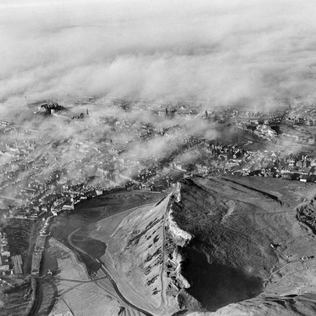 An aerial photo of Holyrood Park. The crags are at the centre of the image and behind them is the city of Edinburgh. The city is under a blanket of smog but here and there church spires and landmarks emerge from the smoke.