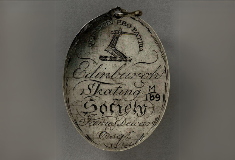 A silver medal engraved with the words Edinburgh Skating Society, James Dewar Esq. The script is very fancy with lots of scrolls. At the top is a representation of an arm holding a sword or dagger and the inscription 'QUIT NON PRO PATRIA'.