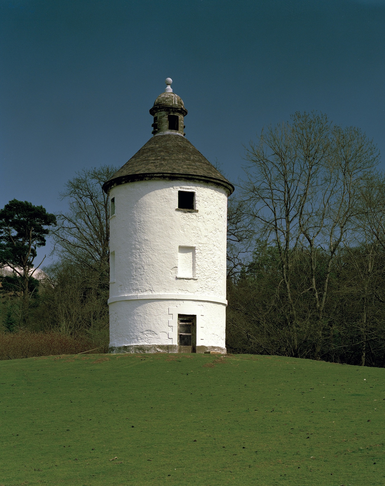 A round white doocot with a dark roof.It has two small windows (one blocked) and a small door.