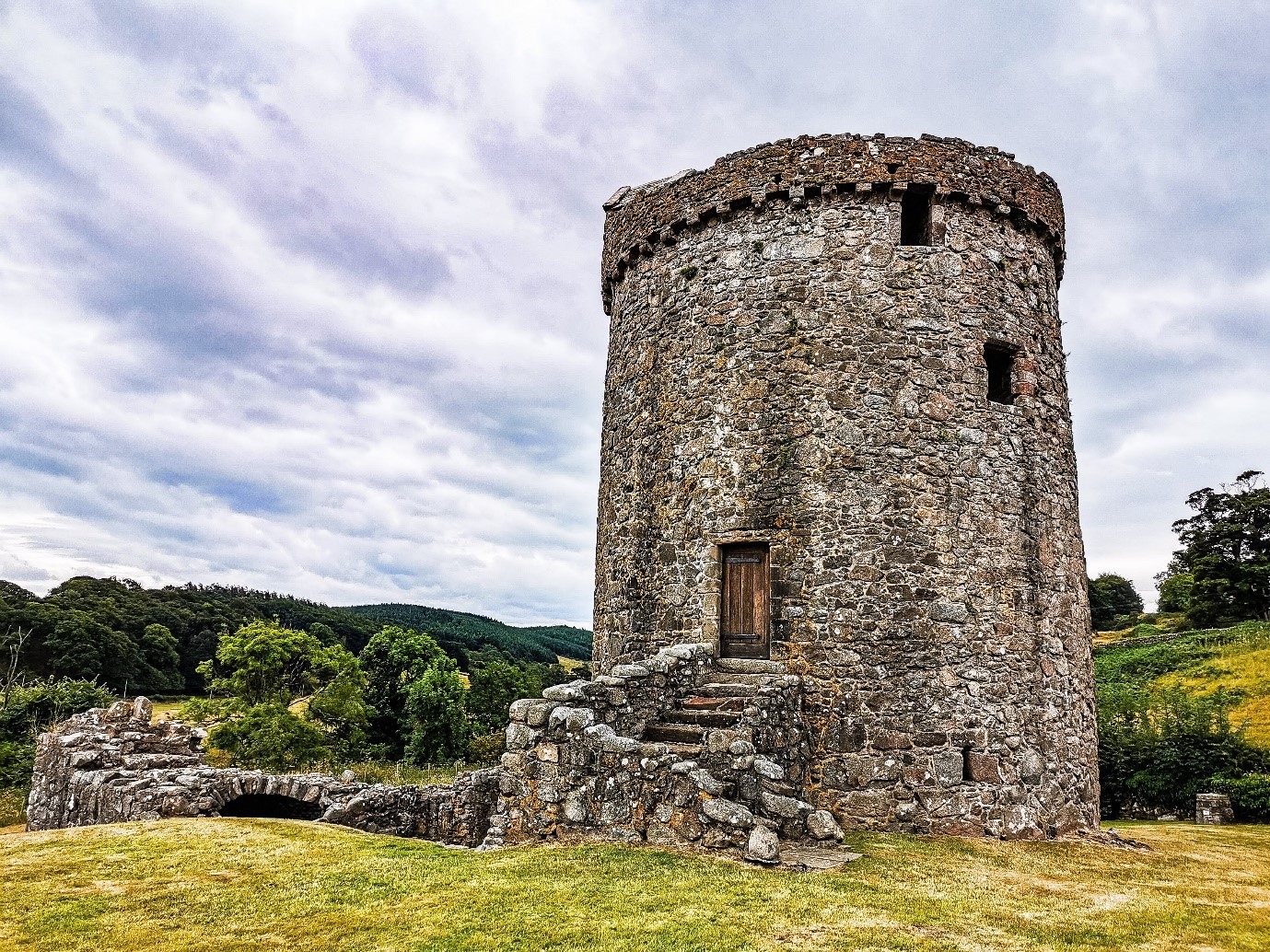 A photograph of Orchardton Tower from the front. It is a cloudy day with blueskys, and trees int he background. The tower is stone with stairs going up it, and a wooden door.