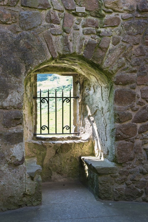 A photo from inside Orchardton Tower, of a window with a black gate. The walls are stone, it is sunny outside with greenery.