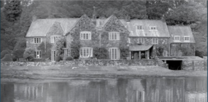 Photograph of the House on the Shore. The photo is in black and white, the house is large with water below it, and trees surrounding it.