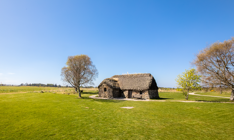 A small, thatched, stone built cottage sits in the middle of Culloden moor. It's a bright sunny day and the sky is blue. The cottage has a bench in front of it and there are a few trees scattered around it.