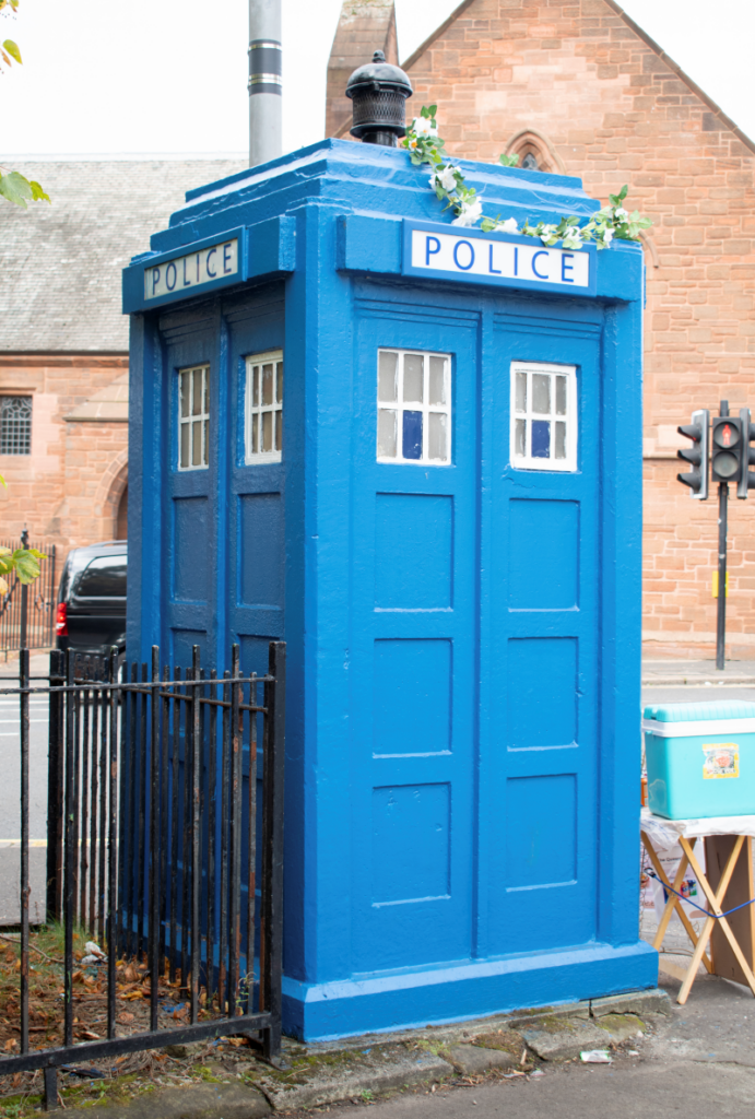 A photograph of a blue police box with flowers on the top of it.
