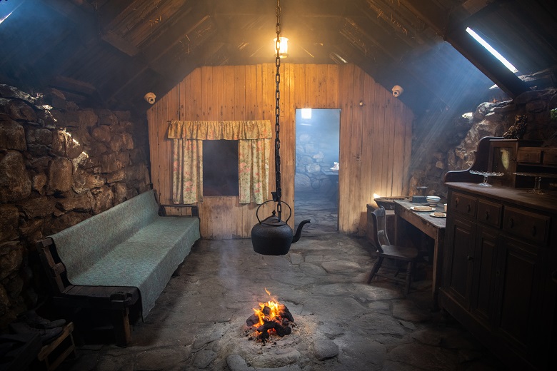 A cosy historic interior of a blackhouse cottage. A large black kettle hangs on a chain from the ceiling over a fire in the middle of the floor. Simple furniture lines the walls of the room.
