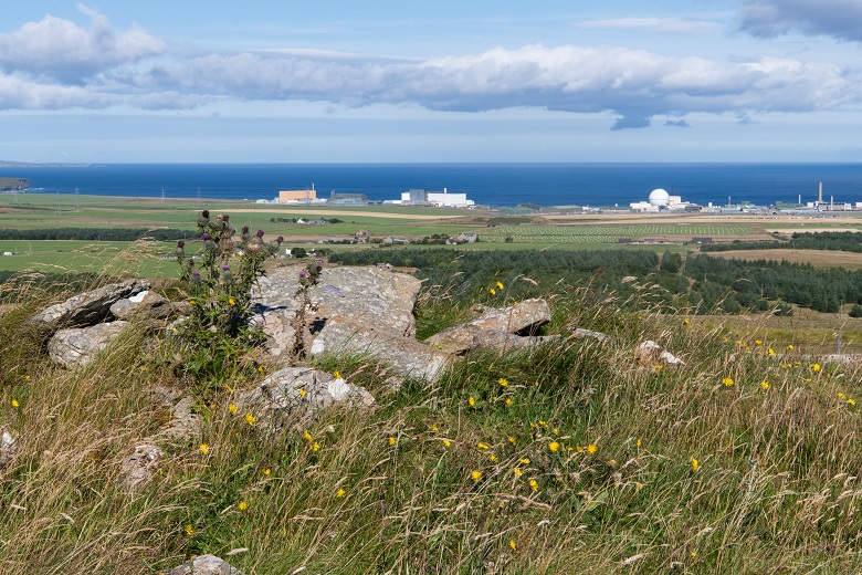 in the foreground a rough pile of large stones on a hillside. In the distance is the sea with a group of industrial looking buildings on the shore.