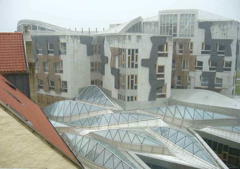 A rooftop view of the Scottish Parliament, featuring several distinctive, leaf-shaped glass domes forming the entrance roof, with the MSP office building rising behind.