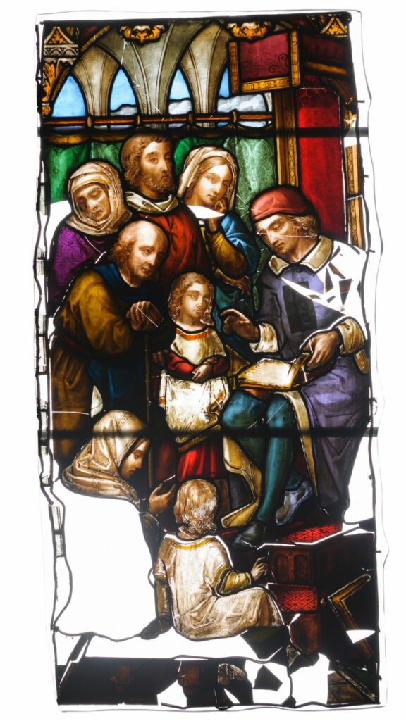 Stained glass. A group of children, men and women gather round a man wearing a red cap, possibly a Cardinal, as he reads to them from a holy book.