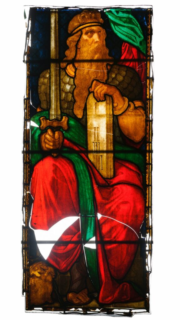 Stained glass of a fierce looking man with a long beard. He is seated and holds a tablet in one hand and a large, upright sword in the other. He wears a red robe with a green cloak draped over him.