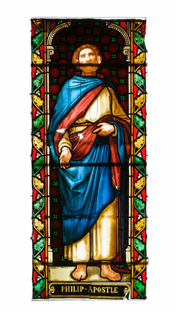 Stained glass showing Philip the Apostle. He wears a white robe with a blue and red cloak thrown over his shoulder. He is bare foot. He is bearded and looking up to the sky.