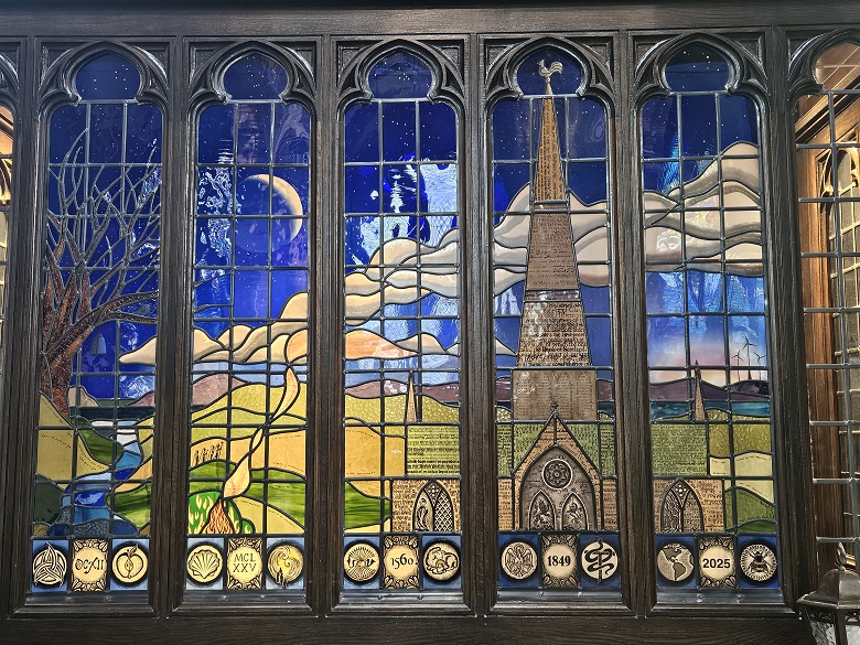 Stained glass window depicting a tall church steeple against a night sky with a crescent moon and rolling hills.