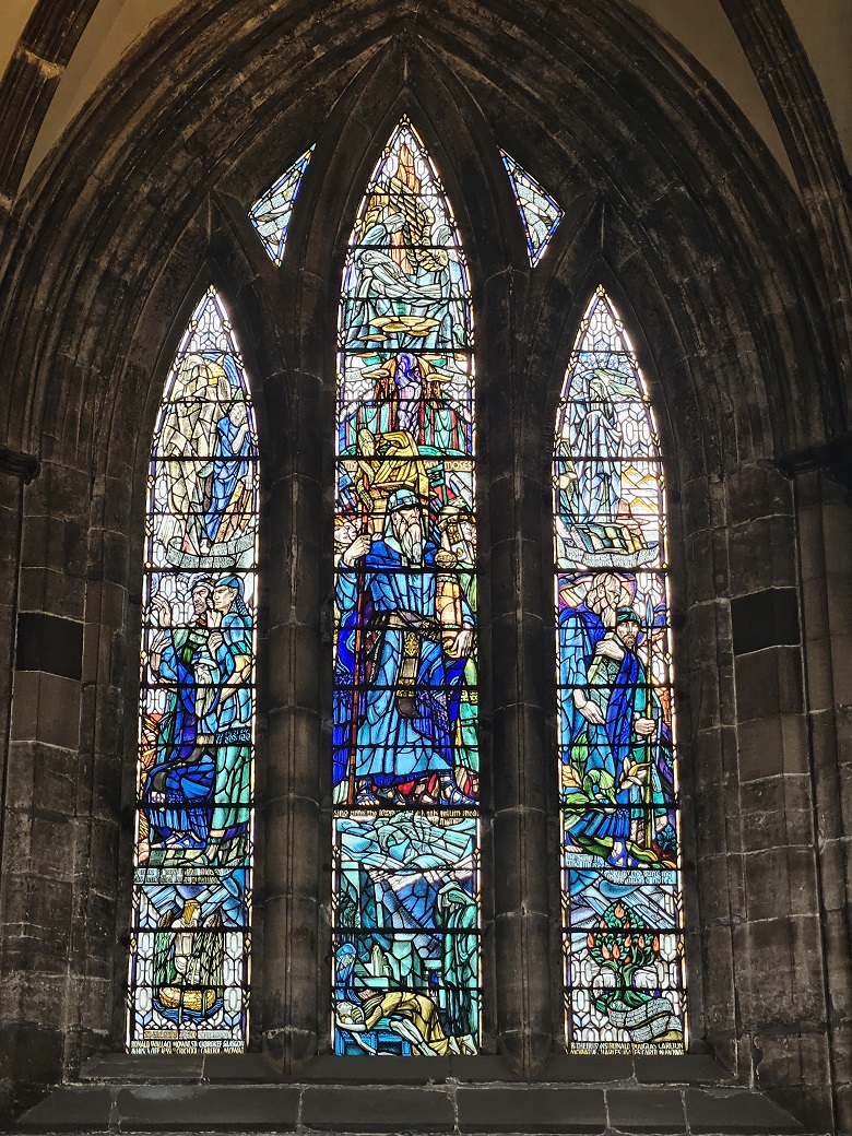 Three tall Gothic stained glass windows depicting biblical scenes with figures in robes and symbolic elements. Moses stands in the middle with a staff and a scroll. Blue and teal are the dominating colours.