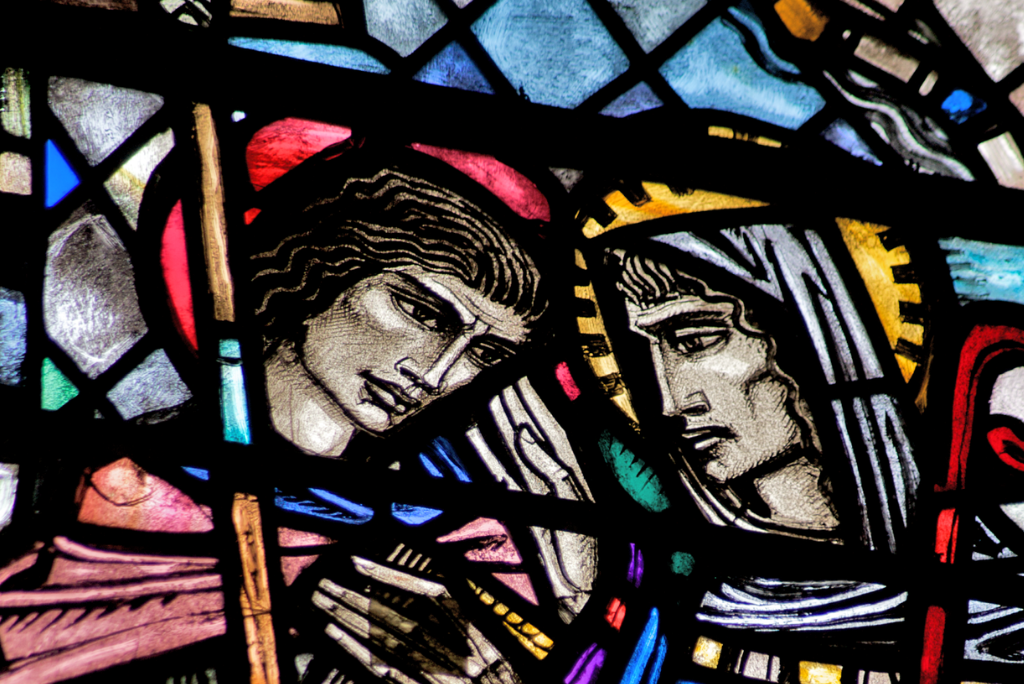 Stained glass depiction of two saints leaning their heads together in a arts and crafts style.