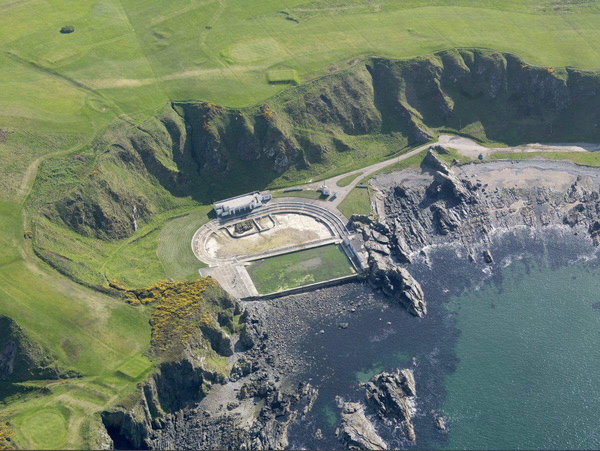 Aerial image in colour showing a rugged landscape surrounding the curved swimming pool in Tarlair