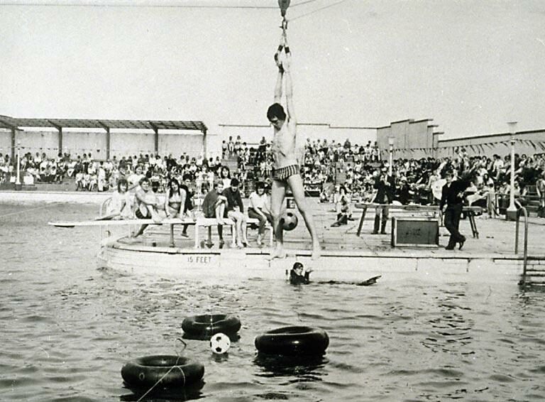 A black and white photo of a person holding on to a hook swinging over the open air pool basin surrounded by crowds.