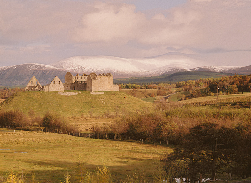 A roofless, stone barracks building perched on top of a grassy hill, with snow capped mountains in the background.