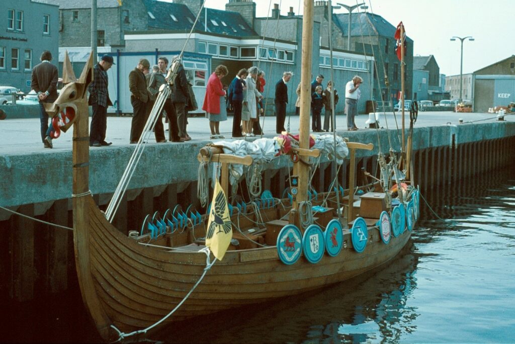 A Viking boat is parked on a river, it is wooden, with blue circles on the side. People are looking at it over a ledge.