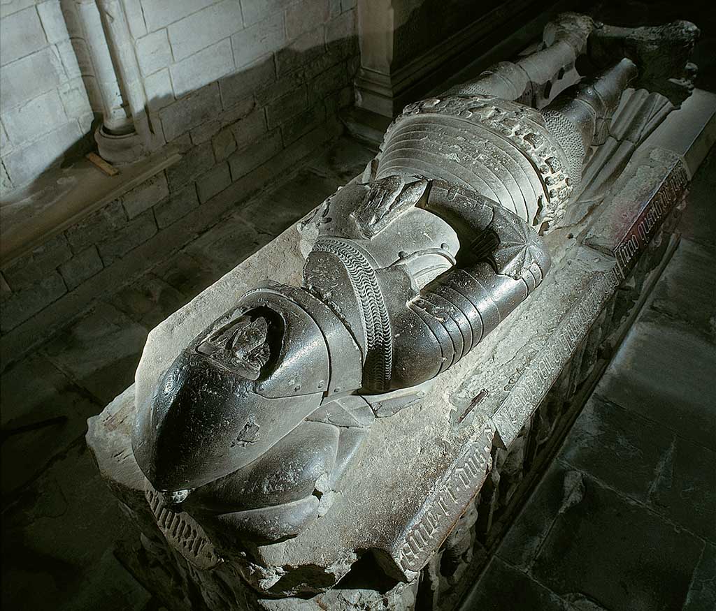 A large stone effigy showing a man clad in full armour and wearing a helmet with his arms folded on his stomach in a prayer position.
