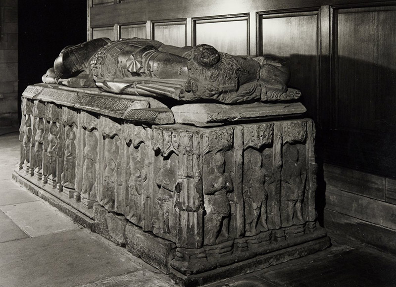 A large stone effigy showing a man clad in full armour. The side of the box is carved with standing warriors