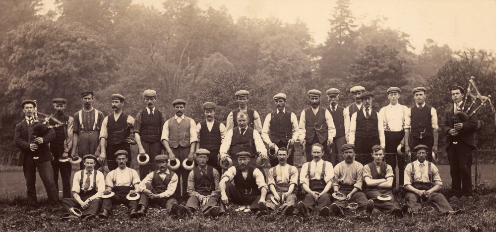 A large group of men in white shirts and waistcoats in two lines, with one row standing and one row sitting in front. Some are holding metal quoits.