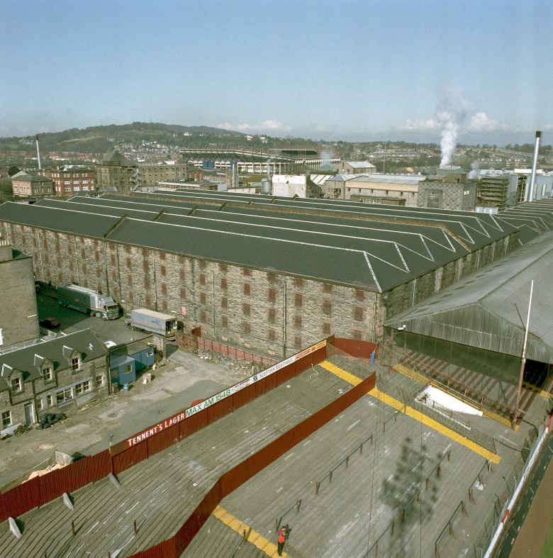 A stadium, with a warehouse next to it, from above. There's a hill in the background.