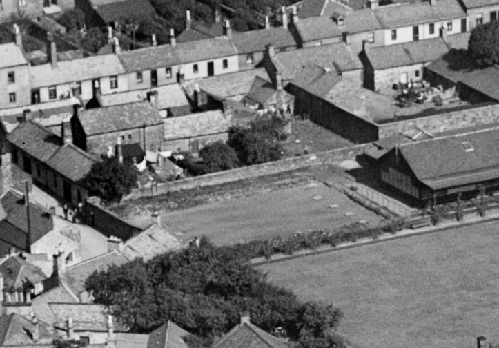 Square grass grounds in the centre of the image, with housing surrounding it, as well as an adjoining field in front. 