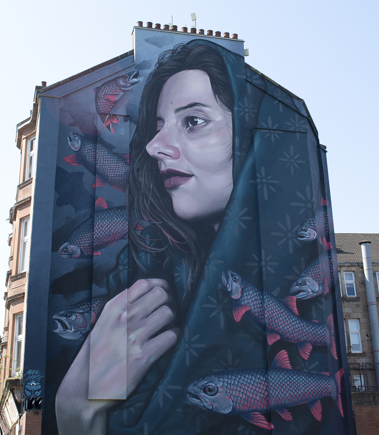 Large mural on the side of a building showing a woman clasping a hooded cloak around herself. Surrounding her are several fish with red fins and scales, swimming against a dark blue background with subtle floral patterns. 