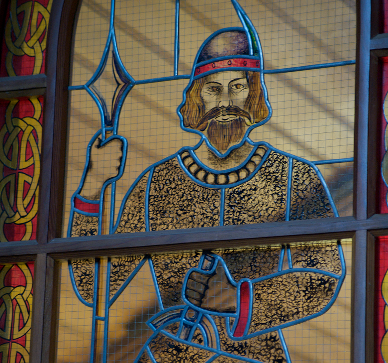 Stained glass window showing a medieval king in ornate armour, holding a spear in the left hand and a sword in the right. The knight wears a helmet. The window is framed with decorative red and gold patterns.