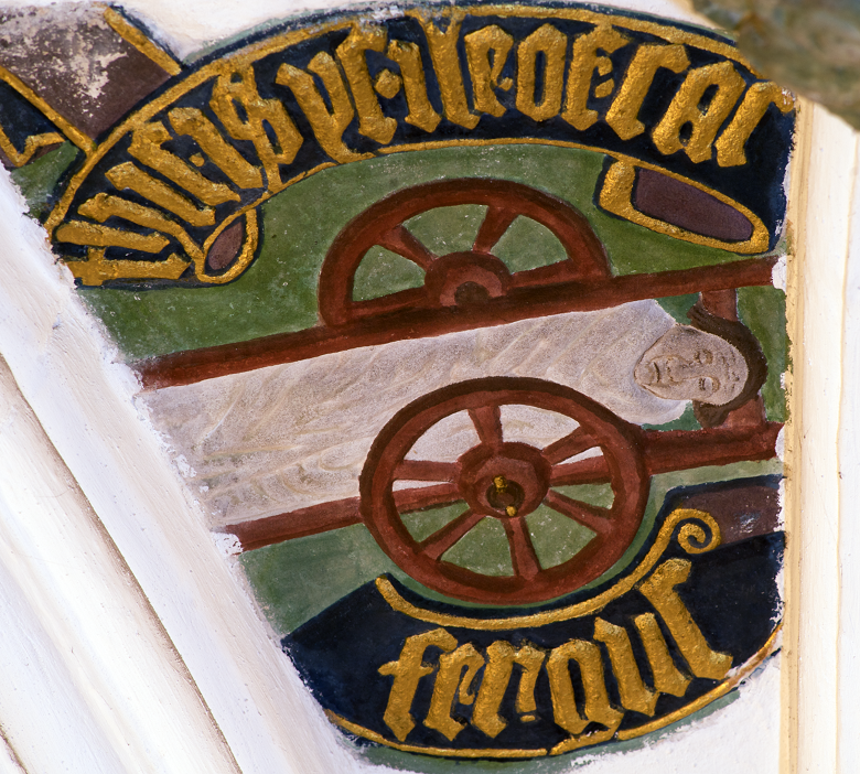 Painted ceiling boss depicting the dead body of St Fergus lying on a cart. The figure is shown with closed eyes and draped in a shroud, surrounded by stylized decorative elements typical of ecclesiastical artwork.