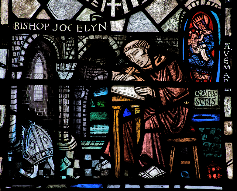 Stained glass window depicting Bishop Jocelyn seated at a desk, writing in a book with a quill. He wears traditional clerical robes. To the left is an ornate archway with a mitre on the floor. To the right, Mary holds baby Jesus, accompanied by the inscriptions “AVE MARIA” and “ORA PRO NOBIS.” The background features intricate architectural details and vibrant stained glass patterns.