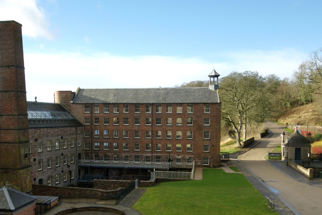 A large, historic brick mill building with multiple windows and a tall chimney stands in the background. In front of the building is a well-maintained green lawn and a circular stone structure. A road runs alongside the building, leading to a gatehouse and trees in the distance under a clear blue sky.