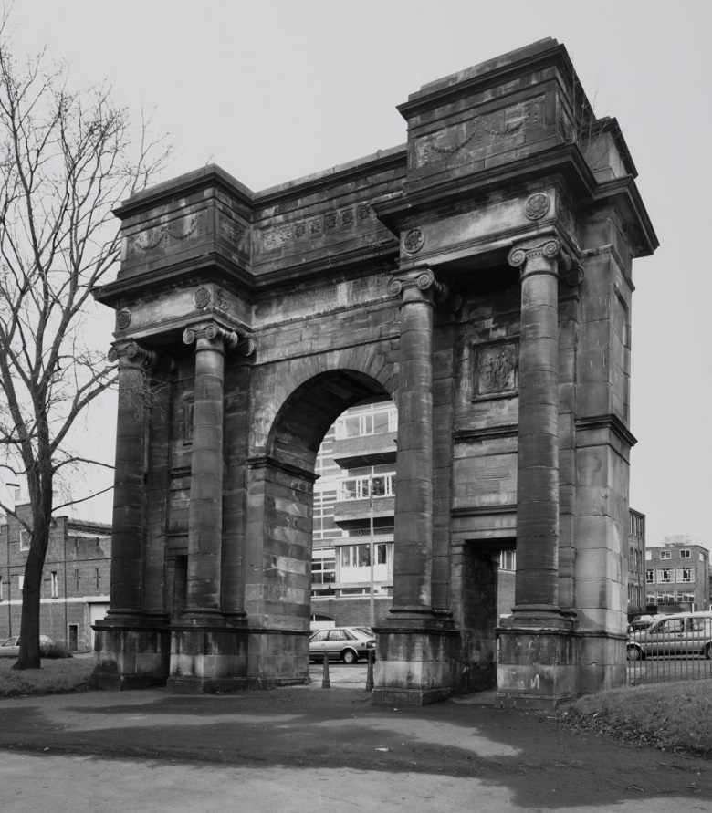 Black and white photograph of a historic stone archway flanked by two pairs of tall columns. The arch features intricate carvings and decorative elements at the top. In the background are modern buildings, parked cars, and a leafless tree to the left.