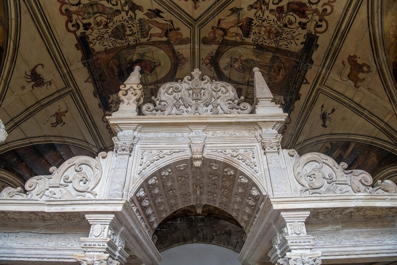 Intricately carved stone archway within a historic church. The arch is flanked by two pointed spires and decorated with ornate scrollwork. Above the arch, the ceiling features frescoes depicting various scenes and figures, showcasing detailed craftsmanship and historical artistry.