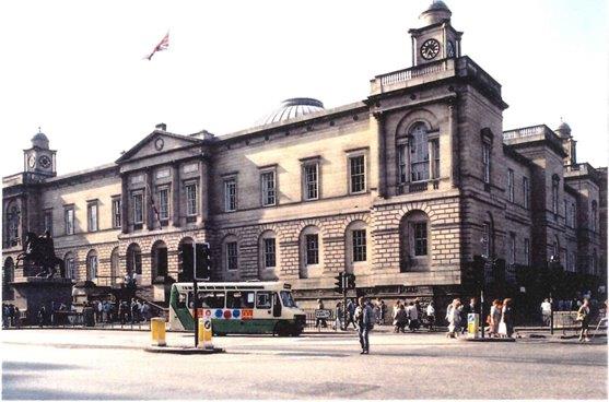 A massive golden stone neoclassical building on the far side of a wide street with people and traffic. There is a flag on a pole in the middle and a large equestrian statue on a tall stone podium in front.