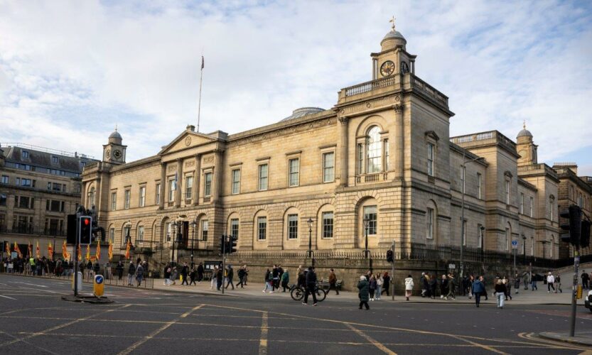 A wide street junction with people walking on a pavement on the far side. Behind them is a large neoclassical stone building. It is two storeys high with a triangular pediment in the centre and high domes towers on two ends.