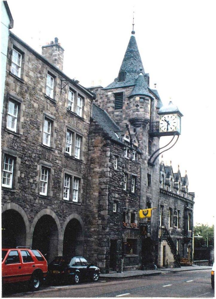 A large rustic stone building with a tall square tower in the middle which has a conical grey slate roof with a metal weather vane on top. A large square clock projects from the tower. There is a road with two cars parked on it in front of the building.