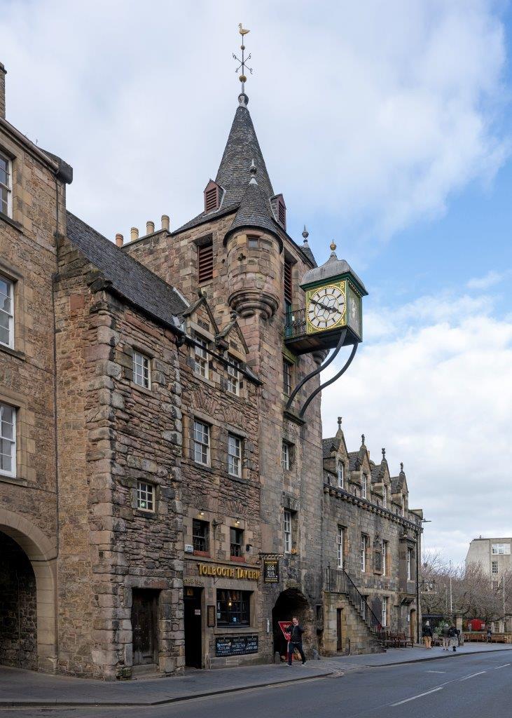 : An empty street with a large stone building on the opposite side, it has a tall tower rising from the middle with a square metal clock jutting from it at the front and a conical slate roof with a weather vane on the top.
