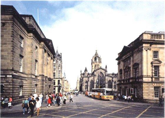 A wide junction of three streets, the middle one stretches into the distance. There are people and a yellow and white coach and large neoclassical buildings to both sides and an impressive church with ornate tower further away.