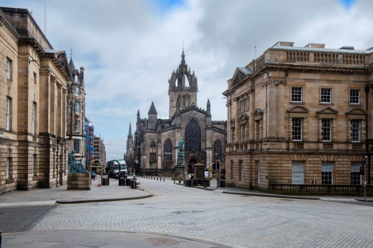 A wide paved street junction with large pale sandstone neoclassical buildings on two corners. A street beyond with a stone gothic church with a high square tower topped by a delicate stone crown.