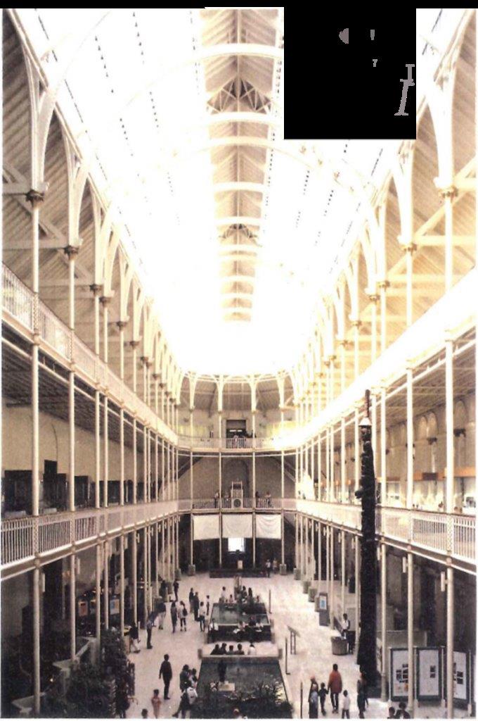 A large three storey room with two deep galleries held up by decorative white cast iron columns around three walls. Light floods from above, the ceiling is high and supported by white iron framework. The floor is pale marble busy with people, two large rectangular ponds and a wooden totem pole.