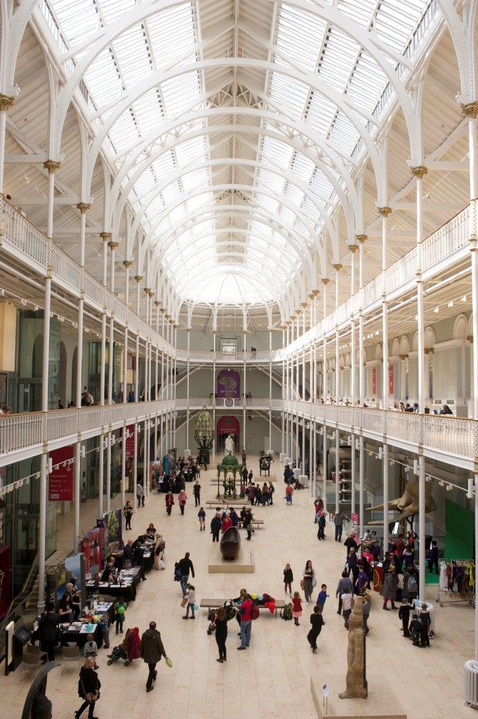 A large three storey room with two deep galleries held up by decorative white cast iron columns around three walls. Light floods from above, the ceiling is high and supported by white iron framework. The floor is pale marble, many people, two large rectangular ponds and a wooden totem pole.
